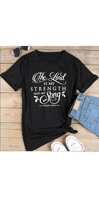 Stylish black t-shirt with inspirational Christian text "The Lord is my strength and song" displayed on wooden backdrop with casual accessories.