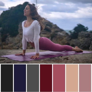 A woman wearing a white top and burgundy leggings practicing yoga outdoors on a yoga mat against a natural landscape.