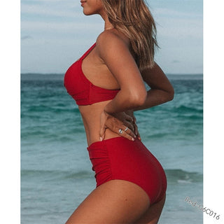 Vibrant red women's beach swimsuit with criss-cross top and high-waisted bottoms, showcasing the model's toned figure against the scenic ocean backdrop.