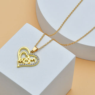 Heart-shaped pendant necklace with "MOM" lettering and diamond accents, displayed on a gold chain against a neutral background.