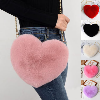 Plush heart-shaped shoulder bag with metallic chain strap, featured in various pastel colors like pink and purple, hanging on woman's jeans.
